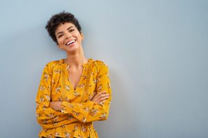 Photograph of a woman with short brown hair smiling with her arms crossed while leaning on a blue wall.