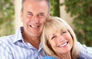 Photograph of an older couple with white teeth holding each other and smiling while looking at the camera.