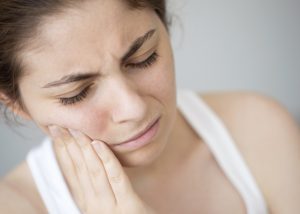 Woman with brown hair tied back and wearing sleeveless white shirt is holding her jaw in pain.