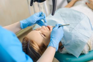 Female with straight dirty blonde hair lying in a dentist chair as a dentist cleans her teeth using two tools.
